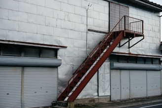 A metal staircase painted in rust-red rises along a large building with a silvery-grey exterior. Below, several closed roller shutters line the building, each in a light grey color. Above the shutters, there are small, rectangular windows with frosted or textured glass. The surface of the building appears to be made from large metal panels, some showing signs of wear or slight dents.