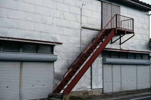 A metal staircase painted in rust-red rises along a large building with a silvery-grey exterior. Below, several closed roller shutters line the building, each in a light grey color. Above the shutters, there are small, rectangular windows with frosted or textured glass. The surface of the building appears to be made from large metal panels, some showing signs of wear or slight dents.
