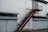 A metal staircase painted in rust-red rises along a large building with a silvery-grey exterior. Below, several closed roller shutters line the building, each in a light grey color. Above the shutters, there are small, rectangular windows with frosted or textured glass. The surface of the building appears to be made from large metal panels, some showing signs of wear or slight dents.