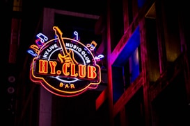A vibrant neon sign with the words 'HY CLUB' in bold letters is featured prominently. It includes musical notes and a guitar graphic, suggesting a music-themed venue. The sign glows in bright colors against a dark background, with architectural elements visible beside it, further enhancing the urban nightlife ambiance.