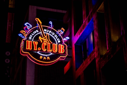 A vibrant neon sign with the words 'HY CLUB' in bold letters is featured prominently. It includes musical notes and a guitar graphic, suggesting a music-themed venue. The sign glows in bright colors against a dark background, with architectural elements visible beside it, further enhancing the urban nightlife ambiance.