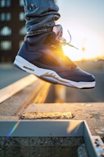 A stylish sneaker with dark blue and white colors is worn by a person wearing ripped jeans. The shoe features a visible air unit and detailed stitching, with a sunset in the background casting a warm glow.