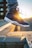 A stylish sneaker with dark blue and white colors is worn by a person wearing ripped jeans. The shoe features a visible air unit and detailed stitching, with a sunset in the background casting a warm glow.