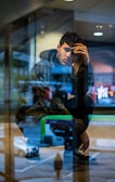 A man crouches by a window, looking pensive and deep in thought. He is wearing a camouflage jacket and dark sneakers. Reflections in the glass add layers to the image, showing parts of the room and the outdoor scene behind him. The ambient lighting creates a contemplative atmosphere.