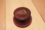 A round, dark red object with a logo featuring a dog and a phonograph is displayed on a light wooden surface. The logo reads 'His Master's Voice' in gold lettering.