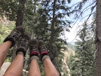 Two pairs of legs with hiking boots and cozy socks are stretched out amidst a landscape of dense, green pine trees. The scene overlooks a mountainous vista under a bright sky, suggesting a peaceful and adventurous outdoor setting.