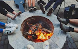 Several people sit around a stone fire pit with a burning fire, each with their legs stretched out towards the center. The scene includes a variety of footwear and an acoustic guitar case is visible nearby, suggesting a casual and relaxed gathering.