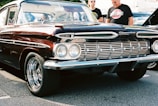 A vintage Chevrolet car with a deep burgundy color and chrome accents is parked on a cement surface. The car features distinct dual headlights and a polished grille, with shiny chrome hubcaps on the wheels. Two individuals stand beside the car, one wearing sunglasses and both dressed casually.