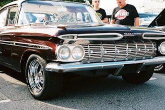 A vintage Chevrolet car with a deep burgundy color and chrome accents is parked on a cement surface. The car features distinct dual headlights and a polished grille, with shiny chrome hubcaps on the wheels. Two individuals stand beside the car, one wearing sunglasses and both dressed casually.