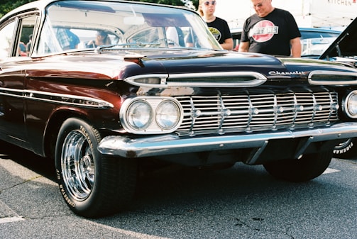 A vintage Chevrolet car with a deep burgundy color and chrome accents is parked on a cement surface. The car features distinct dual headlights and a polished grille, with shiny chrome hubcaps on the wheels. Two individuals stand beside the car, one wearing sunglasses and both dressed casually.