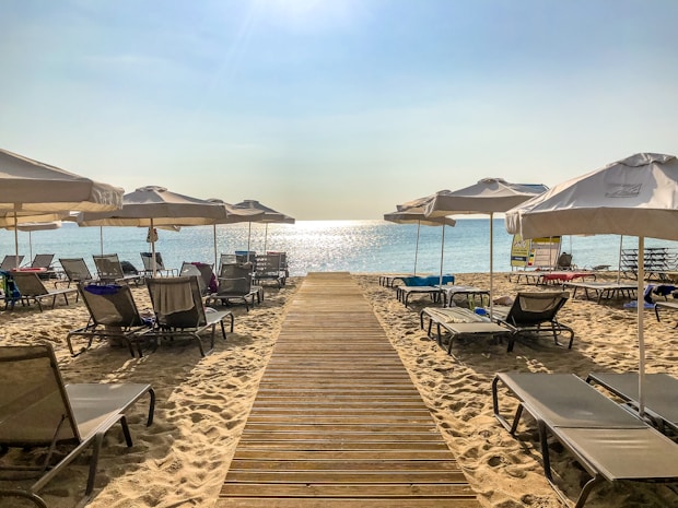 A sunny beach scene with wooden walkways leading to the glistening sea. Multiple beach loungers are neatly aligned under white parasols on the golden sand. The sun is bright, creating a serene and inviting atmosphere.