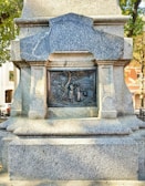 A detailed stone monument displaying an intricate bronze relief set within a rectangular frame. The relief features three figures standing near a tree, with surrounding text and carvings. The monument is situated in an outdoor area, with trees and buildings visible in the background.