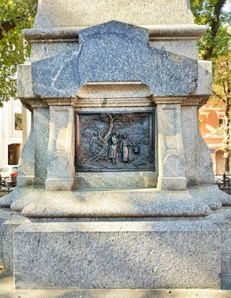 A detailed stone monument displaying an intricate bronze relief set within a rectangular frame. The relief features three figures standing near a tree, with surrounding text and carvings. The monument is situated in an outdoor area, with trees and buildings visible in the background.