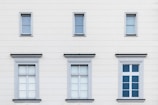 A facade of a building with six windows arranged in two rows. The upper row contains three small windows, each with simple frames, while the lower row features three larger windows with decorative frames. The building's exterior is a light cream color with subtle horizontal lines.