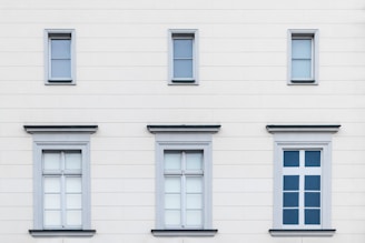 A facade of a building with six windows arranged in two rows. The upper row contains three small windows, each with simple frames, while the lower row features three larger windows with decorative frames. The building's exterior is a light cream color with subtle horizontal lines.