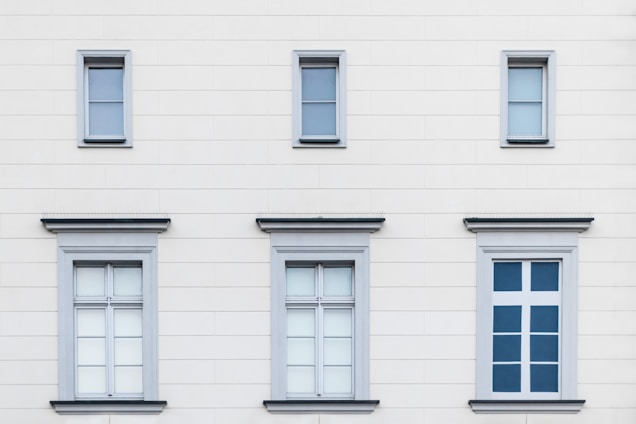 A facade of a building with six windows arranged in two rows. The upper row contains three small windows, each with simple frames, while the lower row features three larger windows with decorative frames. The building's exterior is a light cream color with subtle horizontal lines.