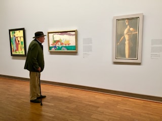 An elderly man with a cane stands in a gallery, wearing a hat and green jacket, observing abstract paintings mounted on a white wall. The paintings are vibrant, with one displaying colorful geometric figures and another showcasing a nude figure.