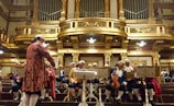 A group of musicians dressed in classical period costumes, including wigs and ornate clothing, are seated in an opulent concert hall featuring elaborate gold architectural details. The musicians are preparing their string instruments, specifically violins and violas, with sheet music in front of them. A conductor in similar period attire is standing in front of the group.