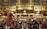 A group of musicians dressed in classical period costumes, including wigs and ornate clothing, are seated in an opulent concert hall featuring elaborate gold architectural details. The musicians are preparing their string instruments, specifically violins and violas, with sheet music in front of them. A conductor in similar period attire is standing in front of the group.