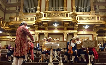 A group of musicians dressed in classical period costumes, including wigs and ornate clothing, are seated in an opulent concert hall featuring elaborate gold architectural details. The musicians are preparing their string instruments, specifically violins and violas, with sheet music in front of them. A conductor in similar period attire is standing in front of the group.