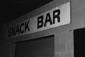 A black and white photograph of a building exterior with a sign reading 'SNACK BAR'. The sign is prominently displayed on a brick wall above a closed metal shutter or garage door. The overall appearance is somewhat aged, and the lighting suggests it may be night or in a low-light setting.