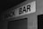 A black and white photograph of a building exterior with a sign reading 'SNACK BAR'. The sign is prominently displayed on a brick wall above a closed metal shutter or garage door. The overall appearance is somewhat aged, and the lighting suggests it may be night or in a low-light setting.