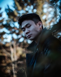 A person is standing in a natural outdoor setting, surrounded by blurred trees and foliage. The lighting is soft and the colors are muted, creating a calm and introspective atmosphere. The person is wearing a dark coat and has a gentle, contemplative expression.