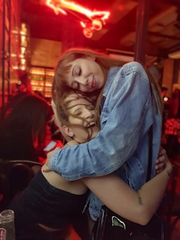 Two people are embracing affectionately in what appears to be a dimly lit bar or restaurant. They are smiling, with one wearing a denim jacket and the other in a sleeveless top. Red lighting creates a warm and intimate atmosphere around them.