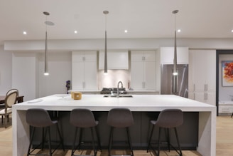 A modern kitchen features a large white marble island with four gray bar stools. The kitchen is equipped with stainless steel appliances and white cabinetry. Pendant lights hang above the island, and there is a colorful abstract painting on the wall.
