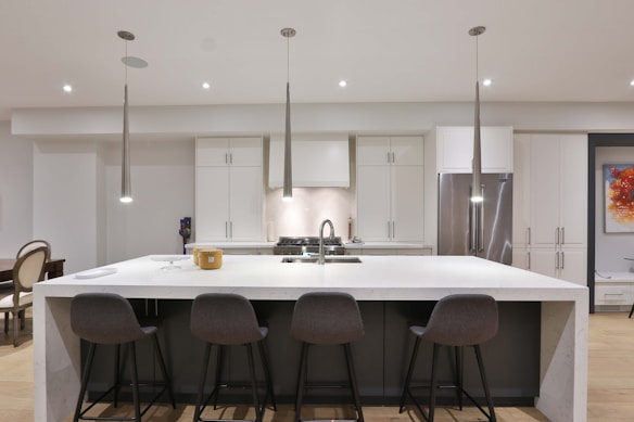A modern kitchen features a large white marble island with four gray bar stools. The kitchen is equipped with stainless steel appliances and white cabinetry. Pendant lights hang above the island, and there is a colorful abstract painting on the wall.
