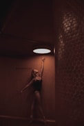 A dancer in a dark leotard gracefully stretches upward, reaching toward a bright circular light on the ceiling. The setting features warm, textured walls with a word 'Southbound' visible on one side.