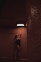A dancer in a dark leotard gracefully stretches upward, reaching toward a bright circular light on the ceiling. The setting features warm, textured walls with a word 'Southbound' visible on one side.