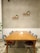 A minimalist room features a long wooden table surrounded by chairs, placed on a concrete floor. Two shelves with small potted plants and decorative items are mounted on the plain, light gray tiled wall. A tissue box and a small object sit at the center of the table.