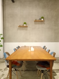 A minimalist room features a long wooden table surrounded by chairs, placed on a concrete floor. Two shelves with small potted plants and decorative items are mounted on the plain, light gray tiled wall. A tissue box and a small object sit at the center of the table.