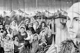 A monochromatic mural depicts a group of people, including women and children, with determined expressions and clenched fists. The backdrop features mountains and traditional houses. A large face of a woman on the right side emphasizes strength and resilience.