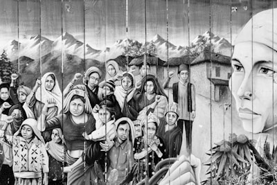 A monochromatic mural depicts a group of people, including women and children, with determined expressions and clenched fists. The backdrop features mountains and traditional houses. A large face of a woman on the right side emphasizes strength and resilience.