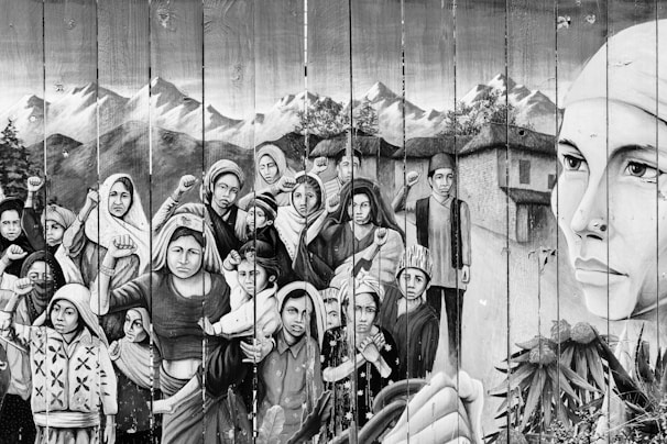 A monochromatic mural depicts a group of people, including women and children, with determined expressions and clenched fists. The backdrop features mountains and traditional houses. A large face of a woman on the right side emphasizes strength and resilience.