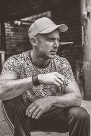 A person wearing a patterned shirt and cap is sitting down, holding a cigarette. The background features a rustic setting with wooden structures and brick walls. The image is in black and white.