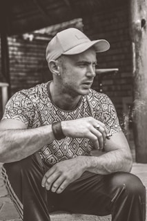 A person wearing a patterned shirt and cap is sitting down, holding a cigarette. The background features a rustic setting with wooden structures and brick walls. The image is in black and white.