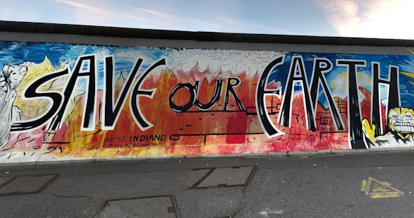 A vibrant street mural features the words 'SAVE OUR EARTH' in large, bold letters. The artwork combines elements of nature and abstract patterns, with clouds and flames depicted in the background. The mural is colorful, featuring shades of blue, red, yellow, and white. It also includes stylized interpretations of natural elements and geometric shapes.