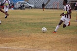 A soccer match is taking place on a grassy field with players in maroon and white striped uniforms. One player is actively kicking a black and white soccer ball, while another player is partially visible. The background shows a sparsely populated seating area in a stadium and a silver car parked nearby.