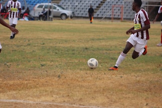 A soccer match is taking place on a grassy field with players in maroon and white striped uniforms. One player is actively kicking a black and white soccer ball, while another player is partially visible. The background shows a sparsely populated seating area in a stadium and a silver car parked nearby.