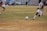 A soccer match is taking place on a grassy field with players in maroon and white striped uniforms. One player is actively kicking a black and white soccer ball, while another player is partially visible. The background shows a sparsely populated seating area in a stadium and a silver car parked nearby.