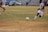 A soccer match is taking place on a grassy field with players in maroon and white striped uniforms. One player is actively kicking a black and white soccer ball, while another player is partially visible. The background shows a sparsely populated seating area in a stadium and a silver car parked nearby.