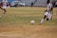 A soccer match is taking place on a grassy field with players in maroon and white striped uniforms. One player is actively kicking a black and white soccer ball, while another player is partially visible. The background shows a sparsely populated seating area in a stadium and a silver car parked nearby.