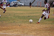 A soccer match is taking place on a grassy field with players in maroon and white striped uniforms. One player is actively kicking a black and white soccer ball, while another player is partially visible. The background shows a sparsely populated seating area in a stadium and a silver car parked nearby.