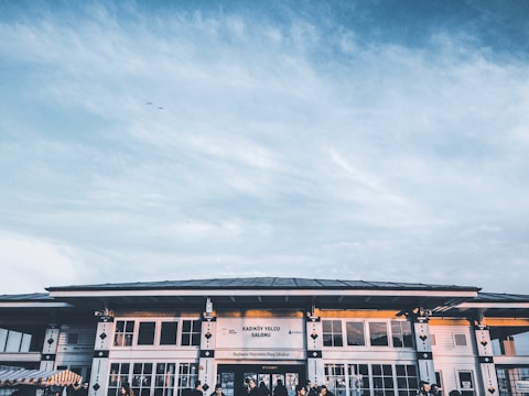 A building with a large sign that reads 'Kadıköy Yolcu Salonu' and additional signage below, against a backdrop of a blue sky with scattered clouds. The structure features large windows with geometric designs and a few figures visible near the entrance.