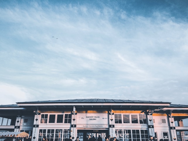 A building with a large sign that reads 'Kadık&ouml;y Yolcu Salonu' and additional signage below, against a backdrop of a blue sky with scattered clouds. The structure features large windows with geometric designs and a few figures visible near the entrance.