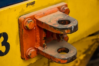 A close-up view of a metal hitch component attached to a painted yellow surface. The hitch is primarily orange with areas showing signs of wear and rust. It is secured with large bolts and has a shiny, worn texture, indicating frequent use.