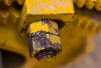 A close-up image of a metal bolt attached to a larger yellow structure, showing signs of rust and wear. The nut is partially threaded over the bolt. The surface is painted in yellow with visible patches of corrosion and chipping paint, surrounded by other metallic parts.